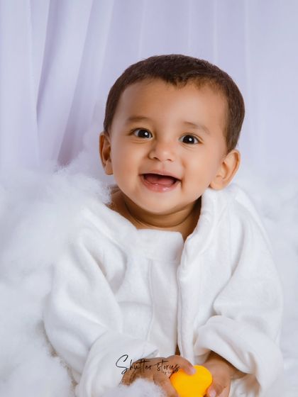 A classic sitter session portrait. This baby boy in a white robe gives a huge, happy smile, sitting in a cloud of white fluff.