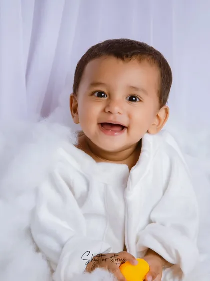 A classic sitter session portrait. This baby boy in a white robe gives a huge, happy smile, sitting in a cloud of white fluff.