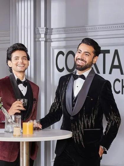 Cocktail chic. Two men enjoying a drink, one in a maroon velvet tuxedo and the other in the black embroidered velvet tuxedo. It's about raising the bar with every detail.