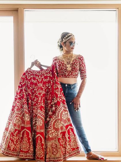 A modern bride with a bit of swag. She pairs her traditional jewelry and blouse with jeans and sunglasses while holding up her intricate red wedding lehenga, showing off her cool and confident personality.