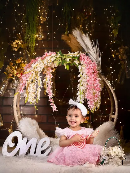 A sweet one-year-old girl in a pink dress, holding a lollipop in our fairy-light and floral hoop setup.