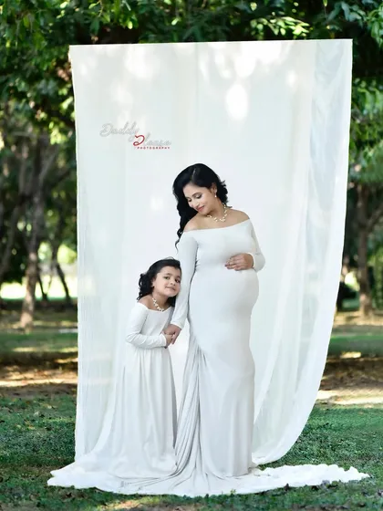 An elegant outdoor maternity shoot with a mother and daughter in matching white gowns. The natural park setting provides a beautiful, serene backdrop for this special moment.