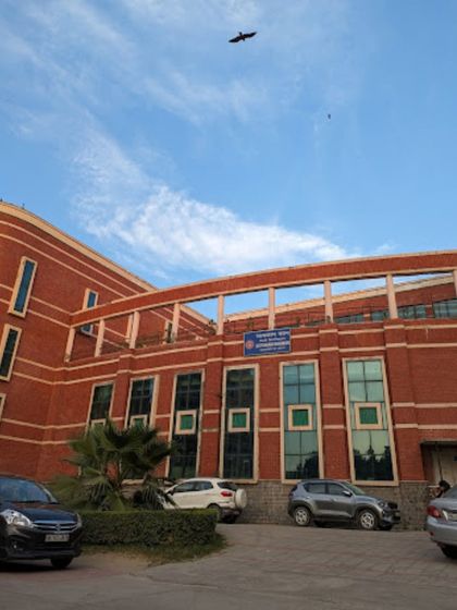A ground-level view of the Delhi University Social Sciences building, showing the interplay of red brick, glass, and the open-to-sky corridor on the upper level, creating a dynamic and functional academic space.