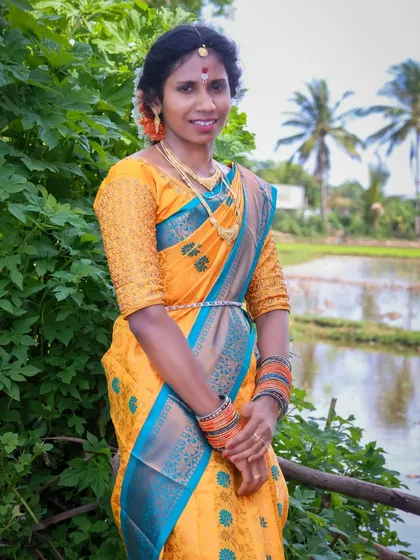 A full-length portrait showcasing a beautiful yellow and blue silk saree. The photo was taken outdoors near a paddy field, adding a touch of local scenery to the traditional look.