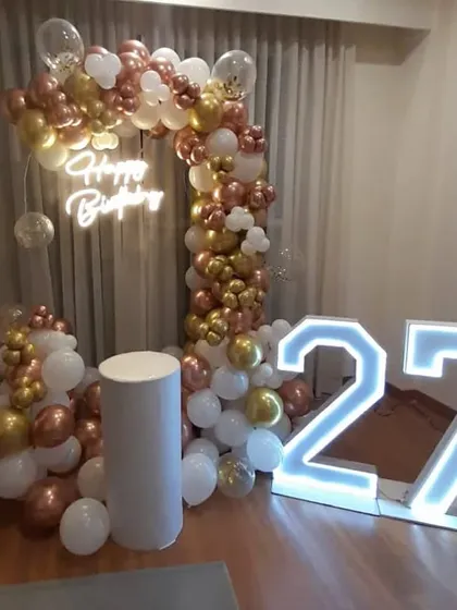 A 27th birthday party with a rose gold, gold, and white balloon arch. The setup includes a "Happy Birthday" neon sign and large marquee numbers.