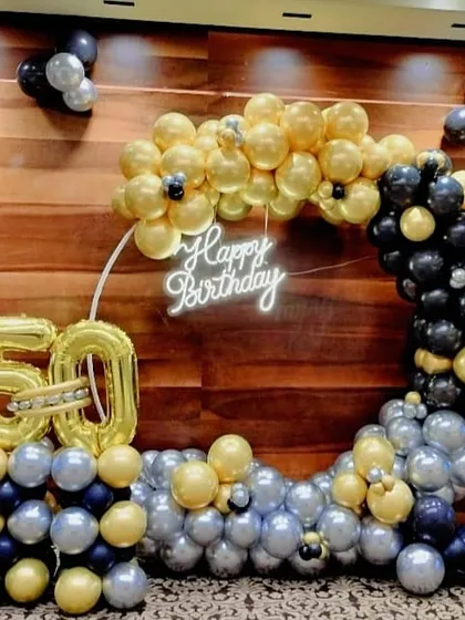 A 50th birthday celebration featuring a half-arch balloon arrangement in gold, black, and silver, with a neon sign and large foil number balloons.