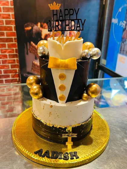 A sharp and stylish two-tier tuxedo cake, perfect for a "Happy Birthday Prince" celebration. The design features a black and white tuxedo with a gold bow tie.
