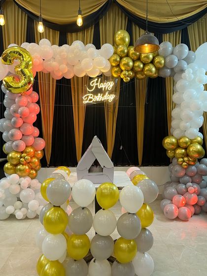 A stylish balloon arch in white, grey, and gold for a 3rd birthday. The setup includes a neon sign and a small teepee, blending kid-friendly and adult aesthetics.