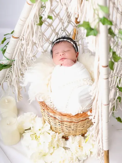 An angelic setup featuring a newborn in a white swaddle, sleeping in a basket inside a boho-style macrame teepee. The white flowers and candles add to the serene atmosphere.