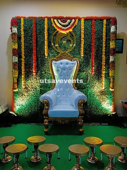 A traditional baby shower backdrop with a grass wall and garlands of fresh flowers. The setup includes a throne chair and brass stands for a classic look.