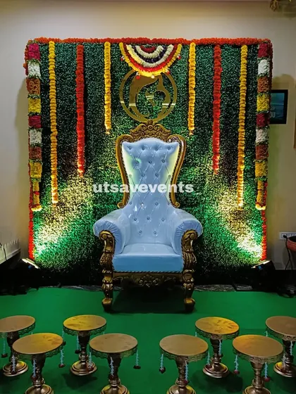 A home baby shower setup featuring a greenery wall with a floral border and a white throne. The lighting from below adds a special glow to the stage.