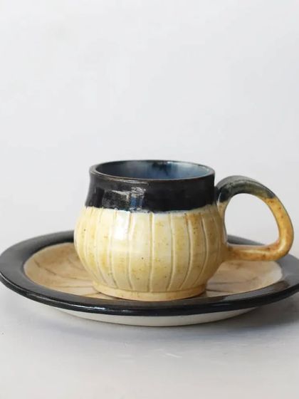 A uniquely shaped coffee cup with a ribbed texture and a two-tone glaze. Paired with a matching saucer, this set is a true conversation starter.