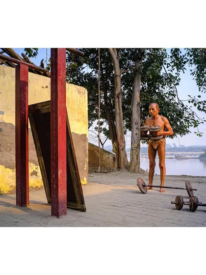 A color photograph of an elderly man lifting weights at an outdoor gym by the river. The warm sunlight and natural setting provide a beautiful backdrop for his workout.