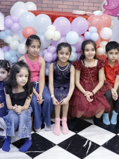 A group of children dressed up for a party, sitting together against a backdrop of festive balloons.