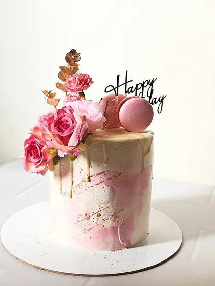A tall, elegant cake with a pink and white watercolor finish, gold drip, and topped with fresh flowers and macarons.