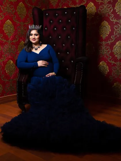 A portrait of power and elegance. The mom-to-be sits on a regal red throne chair, her navy blue gown cascading around her, creating a striking and memorable image.