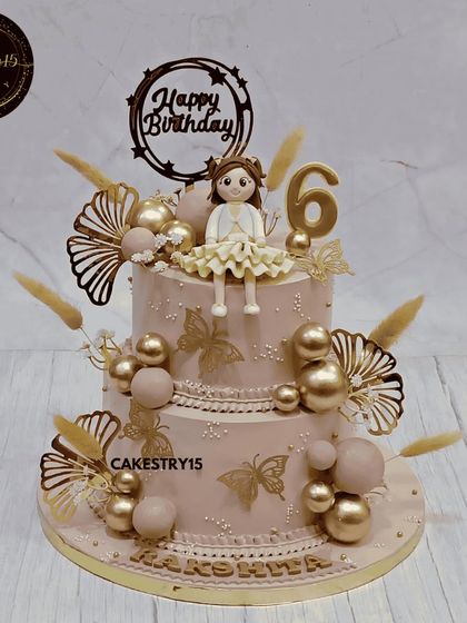 A shimmering and elegant two-tier doll cake. This butterscotch cake is decorated in a dusty rose and gold theme, with a girl figurine, golden butterflies, and delicate dried botanicals for a sophisticated look.