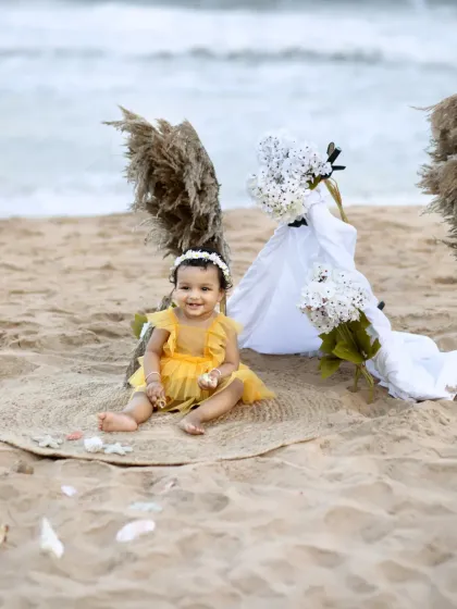 A sweet photo of a baby girl sitting on a jute mat on the beach for her first birthday, with the waves gently rolling in behind her.