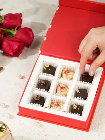 A hand picking a heart-shaped chocolate from a red gift box, surrounded by roses, creating a classic romantic image.
