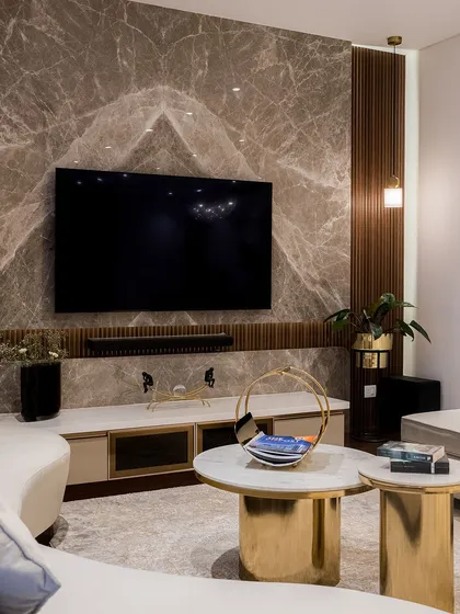 A unique living room design featuring a curved sofa and striking gold coffee tables. The TV is set against a book-matched marble wall, flanked by fluted wood panels, creating a rich textural contrast.