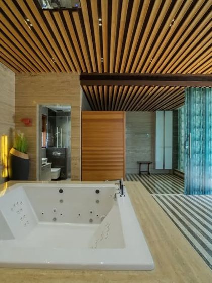 An expansive, luxurious bathroom featuring a large jacuzzi, striped marble flooring, and cast glass partitions. The slatted wood ceiling and warm lighting create a spa-like ambiance.
