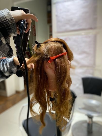 A close-up of a student using a round brush and blow dryer to create volume and curl. Mastering this fundamental skill is essential for any professional hairstylist.