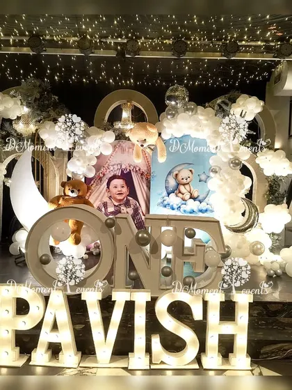 A celestial teddy bear-themed first birthday stage for Pavish. The decor features a crescent moon prop, fluffy cloud balloons, and multiple teddy bears, creating a serene and dreamy atmosphere.