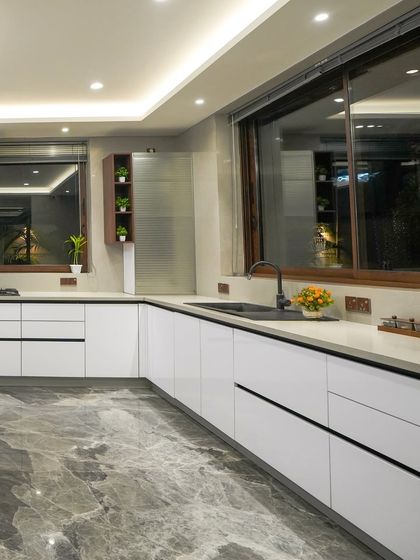 The L-shaped counter layout maximizes workspace, while large windows keep the kitchen bright and connected to the outdoors. The handleless white cabinets maintain a clean, modern aesthetic.