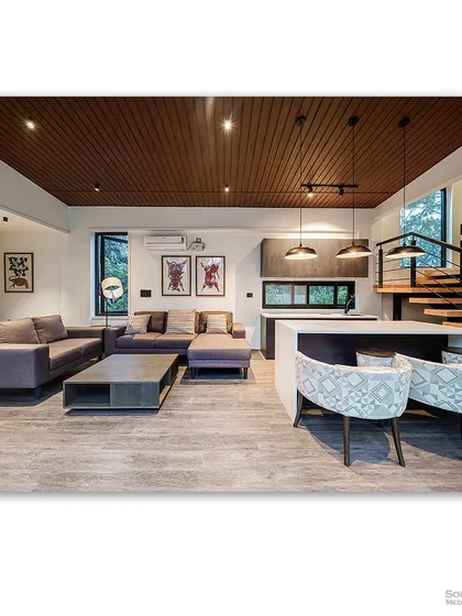 A wide view of the open-plan living area in a container home, featuring a modern kitchen island and a comfortable sectional sofa. The wooden ceiling and grey flooring create a warm yet industrial feel.
