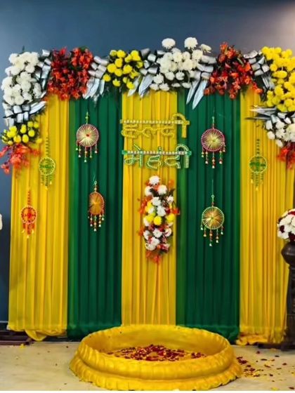 A traditional Haldi and Mehandi setup with a large brass urli. The backdrop features green and yellow drapes, floral arrangements, and dreamcatcher-style hangings.