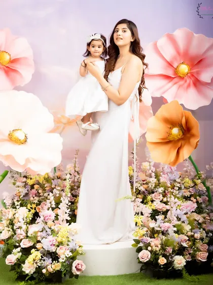 A full-length portrait of a mother and daughter in a beautiful, flower-filled studio setup. Their matching white dresses and the whimsical background create a stunning, high-fashion-inspired image.