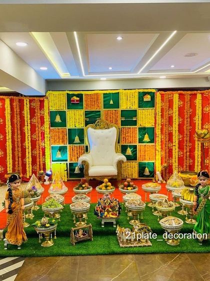A classic and vibrant baby shower decoration with a red, yellow, and green checkered backdrop. The artificial grass flooring and traditional dolls complete the festive look.