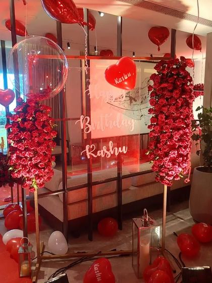 A romantic birthday setup featuring tall pillars of red roses and a personalized acrylic birthday sign.
