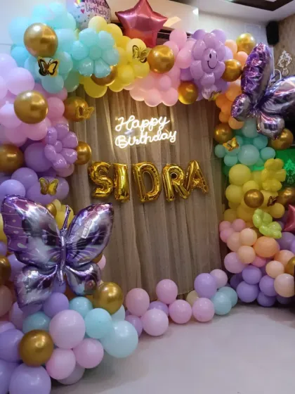 A pastel butterfly-themed decoration with a soft curtain backdrop. The setup includes large butterfly foil balloons and a flower-like balloon arch in shades of purple, pink, yellow, and blue.