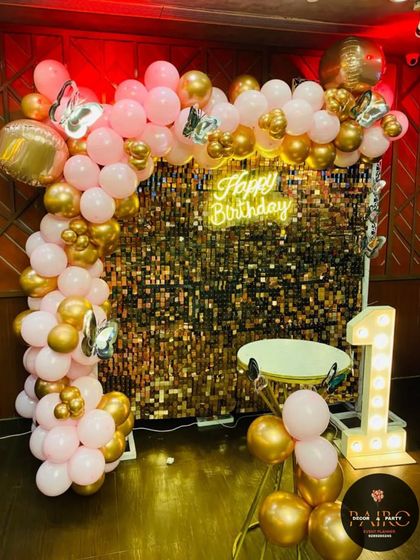 A chic and modern first birthday setup. The decoration is built around a gold shimmer wall, a "Happy Birthday" neon sign, a light-up number "1", and a pink and gold balloon arch.