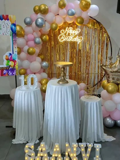 A pink and gold balloon arch with a tinsel backdrop, crown prop, and a photo booth frame.