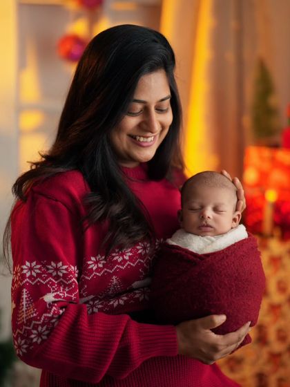 A mother's loving smile as she holds her sleeping newborn. The warm lighting and Christmas decor create a heartfelt, festive atmosphere.