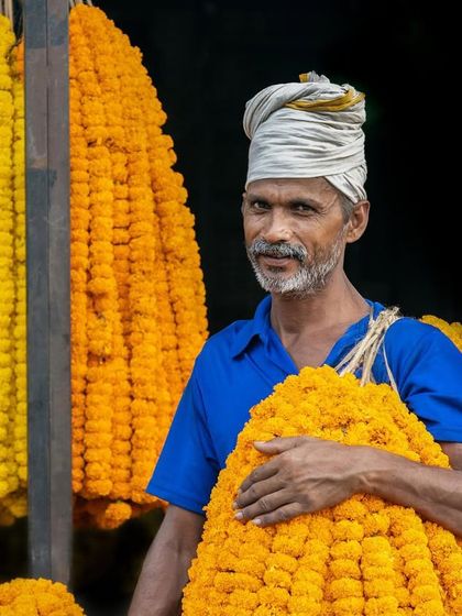 A flower seller in Delhi offers a tired smile, surrounded by vibrant yellow and orange marigold garlands that contrast with his weary expression.