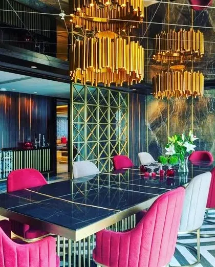 We are drawn to dining rooms that make a bold statement. This space uses dramatic gold chandeliers, a geometric screen, and vibrant pink chairs against a dark, veined marble wall to create an unforgettable, high-energy dining experience.