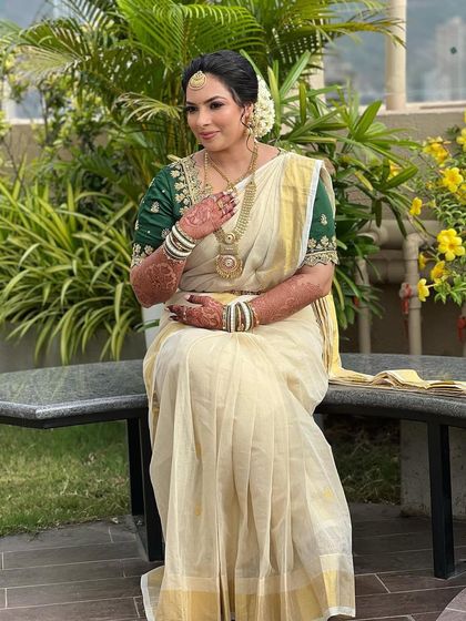 A serene portrait of a Kerala bride in her classic off-white and gold saree, paired with a richly embroidered green blouse. The natural setting highlights her simple elegance.