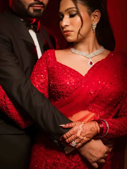 A close-up, intimate portrait focusing on the couple's hands and embrace. The rich red of the bride's saree and the sparkle of her diamond necklace are beautifully highlighted.