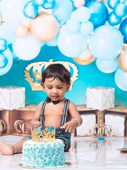 A little prince celebrating his first birthday with a royal blue cake smash. The setup includes a crown, balloons, and a delicious-looking cake ready to be smashed.