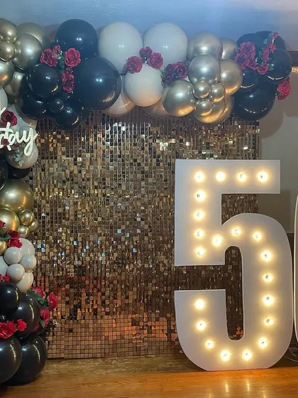 A sophisticated 50th birthday decoration with a gold shimmer wall and a black, gold, and white balloon arrangement. The large marquee numbers make a bold statement.