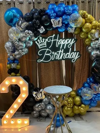 A stylish 2nd birthday party with a circular balloon arch in blue, black, gold, and silver. The setup includes a "Happy Birthday" neon sign and a marquee number '2'.