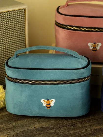 A close-up of Faborange's teal velvet vanity case with its signature bee embroidery. The warm lighting and simple styling draw attention to the product's soft texture and charming details.