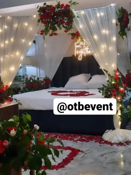 I can turn any bedroom into a romantic escape. This anniversary room decor features a dreamy canopy with sheer drapes and warm fairy lights, surrounded by red roses and a glowing "So Together" neon sign.