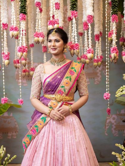 The bride, dressed in a lovely half-saree, poses against the intricate floral backdrop. The hanging garlands of lotus buds and carnations create a beautiful texture and depth for portraits.