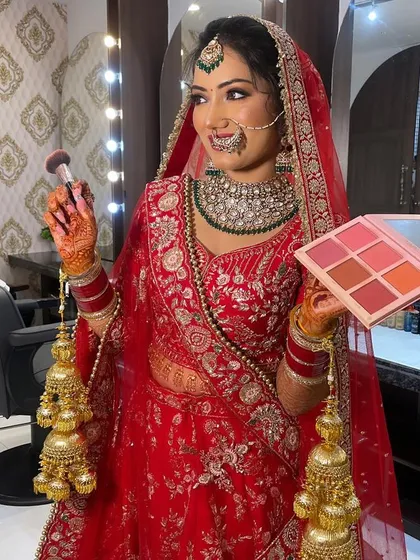 A behind the scenes moment. Our bride is all smiles while getting her final touches, holding a blush palette that complements her red lehenga and green jewelry.