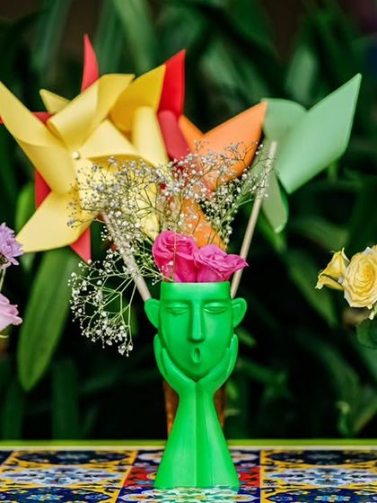 A quirky and colorful centerpiece, featuring a green face-shaped vase holding flowers and pinwheels, set on a table with a vibrant, patterned runner.
