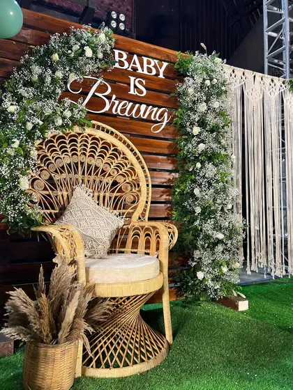 A close-up of the 'Baby is Brewing' boho shower. The focal point is the beautiful peacock chair set against a dark wood wall, framed by baby's breath and a macrame hanging.